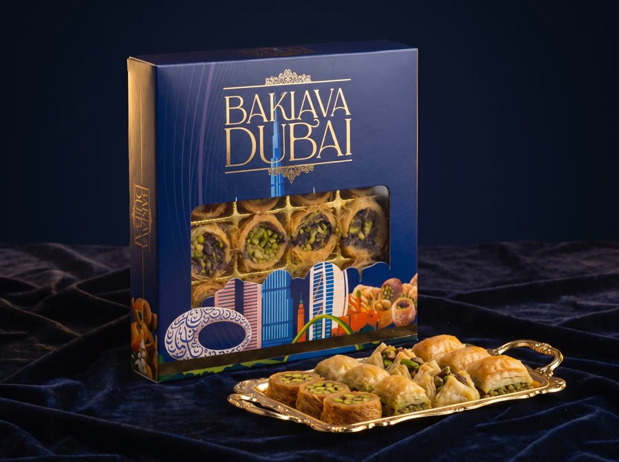 Baklava Dubai gift box by Blue Dubai Sweets with a clear window, beside a gold tray of pistachio baklava