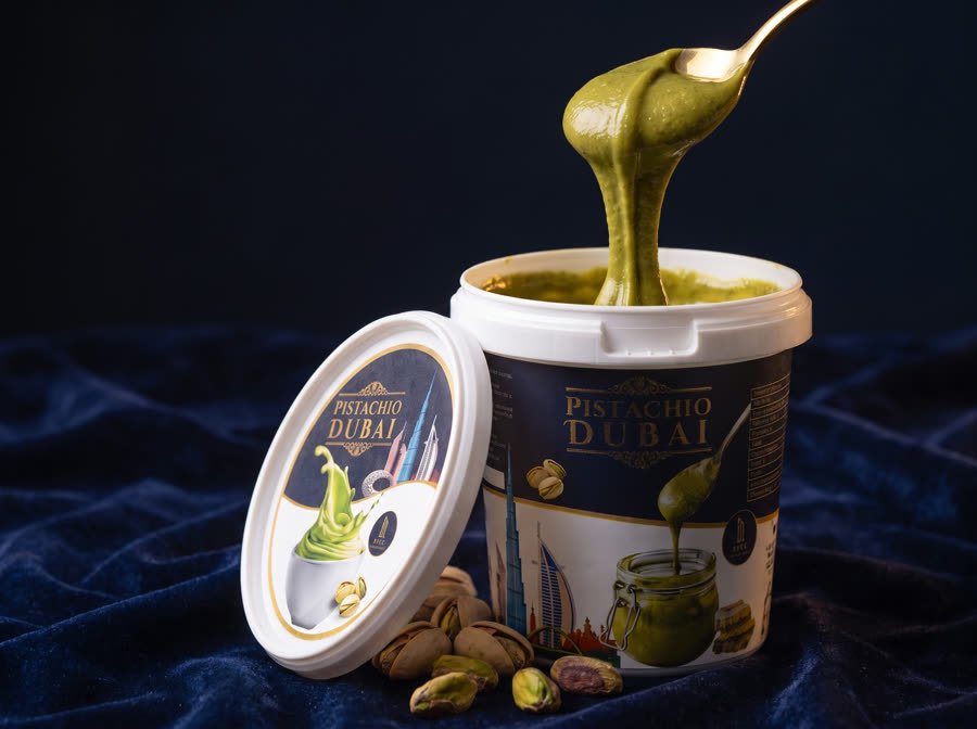 Pistachio Dubai pistachio spread tub by Blue Dubai Sweets with a gold spoon lifting a thick ribbon of green spread